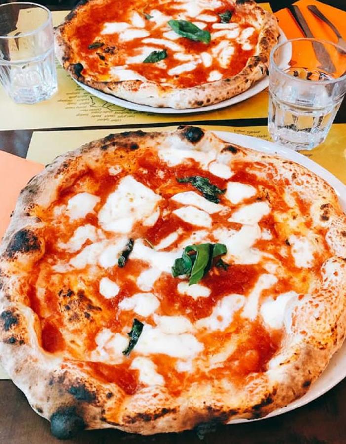 Due pizze Margherita fumanti su tavola, con bicchieri d'acqua e tovaglioli arancioni.